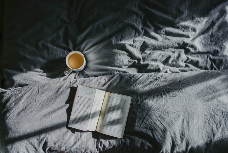 Open book and cup of tea on a bed in soft morning light, symbolising rest and recovery for burnout and performance.
